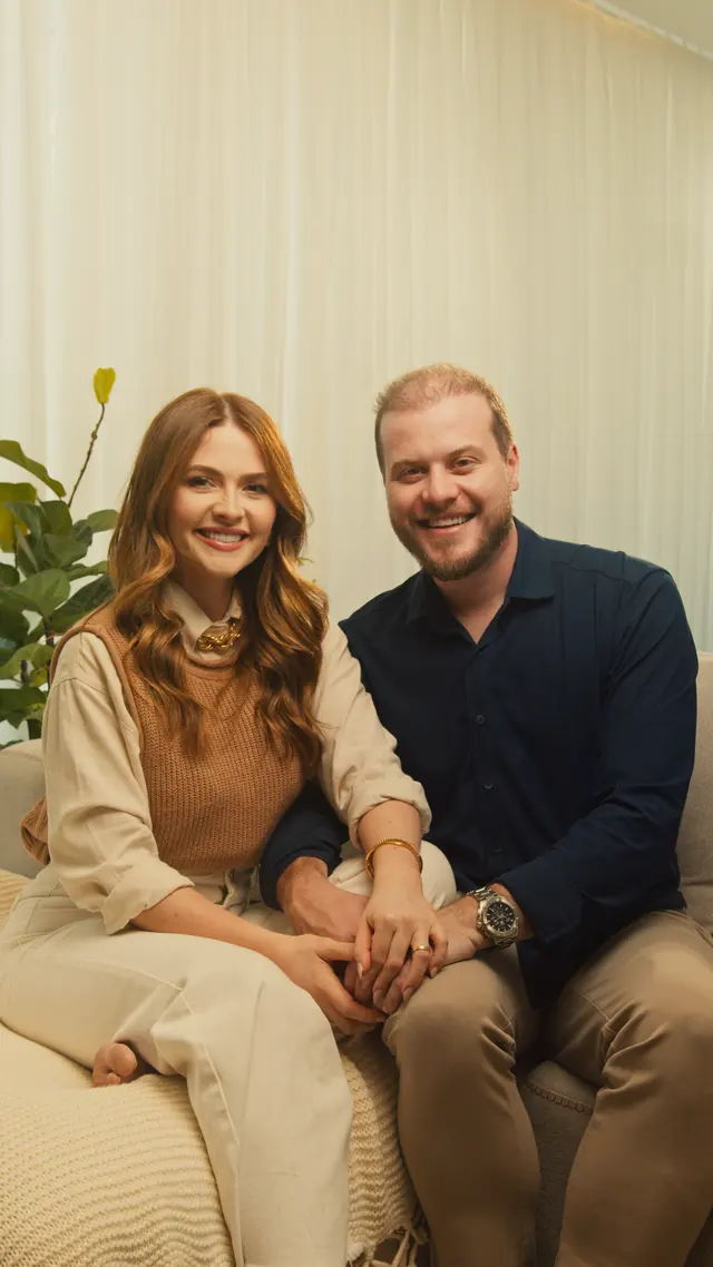 Morgana Santana e o marido sentados lado a lado no sofá, de mãos dadas, sorrindo para a câmera